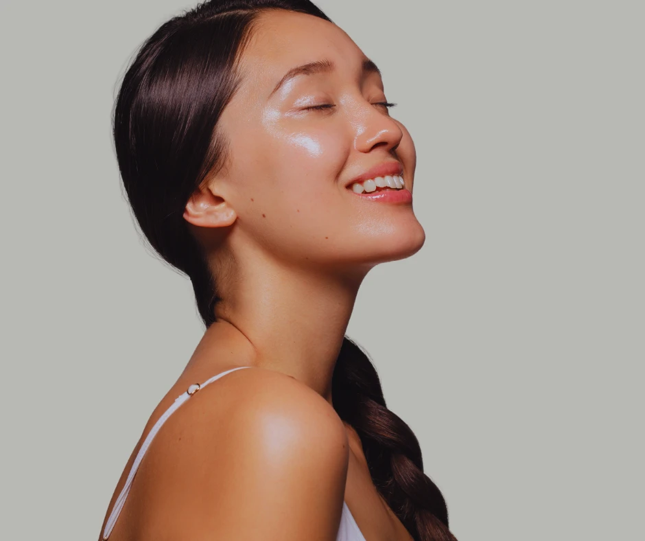 Woman with smooth, glowing skin smiles with her eyes closed against a neutral gray background. Her long dark hair is styled in a loose braid over one shoulder, and she wears a white spaghetti strap top