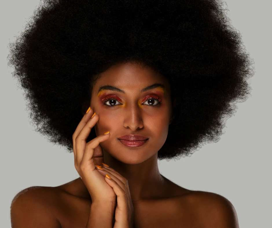 Woman with voluminous natural afro hair poses confidently against a gray background. Her vibrant eye makeup in red and yellow tones complements her glowing skin and gold-tipped nails.