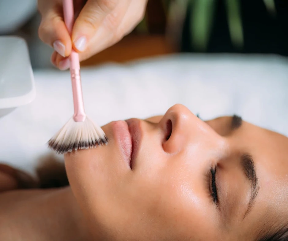 A woman receives a VI Peel facial treatment as a soft brush applies the solution to her glowing skin. She lies relaxed with her eyes closed in a spa setting.