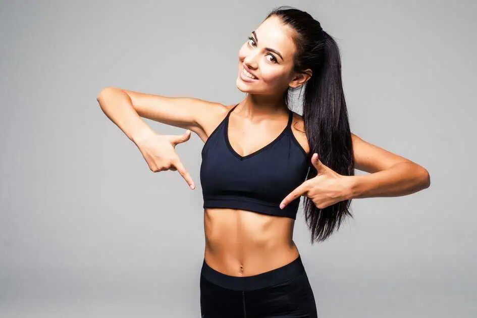 Fit woman in a black sports bra and leggings smiling and pointing at her toned midsection, representing results from mesotherapy lipo dissolve in Houston TX.